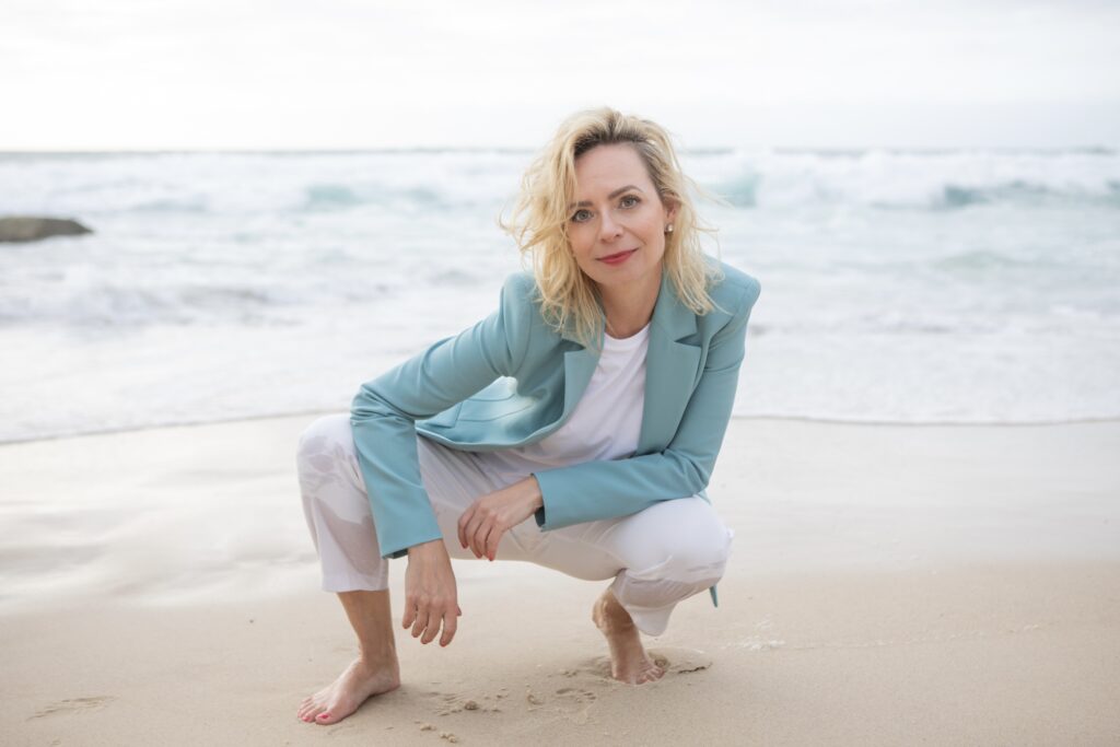 Jenny Heyer hockt barfuß am Strand vor dem Meer.
