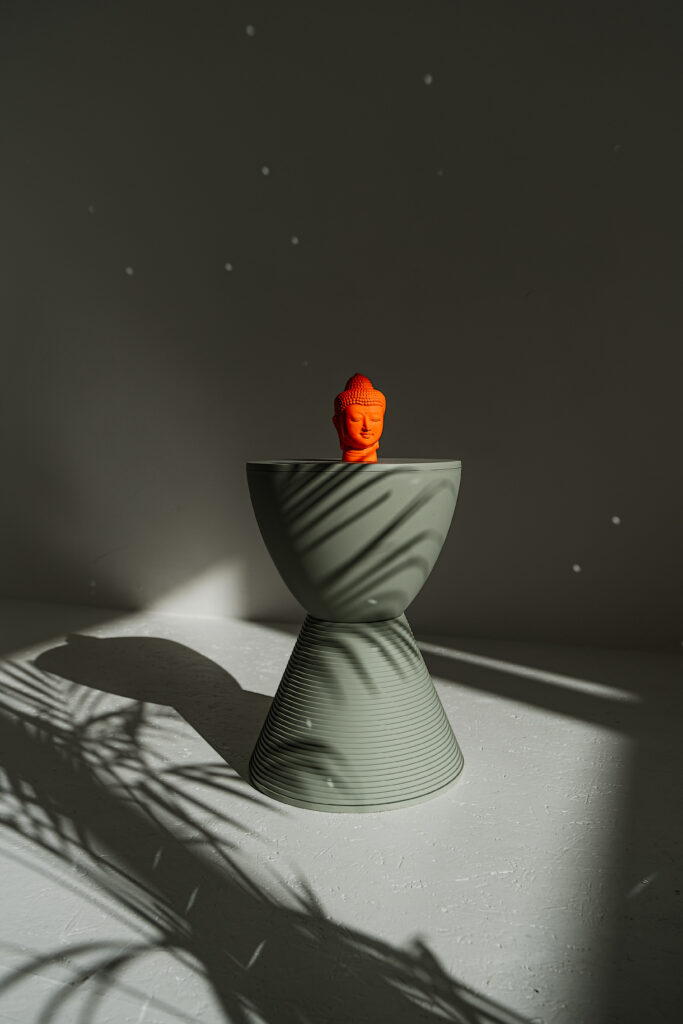 Kleiner orangefarbener Buddha-Kopf auf grauem Hocker im Licht.