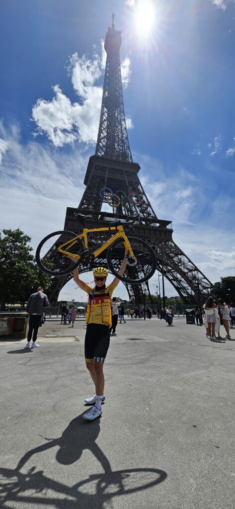 Jenny Heyer in Radkleidung hält ein Fahrrad vor dem Eiffelturm hoch.