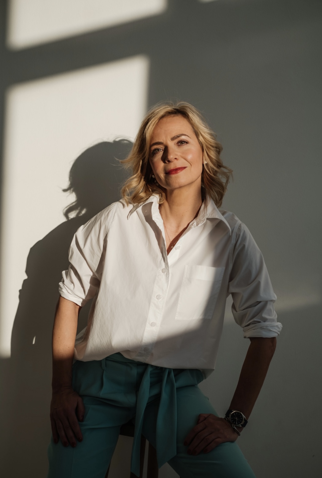 Porträt von Jenny Heyer in weißer Bluse und türkisfarbener Hose vor heller Wand mit Licht und Schatten.