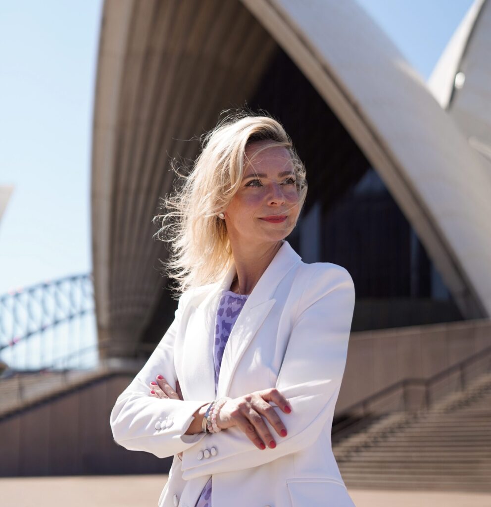 Jenny Heyer in weißem Blazer vor moderner Architektur im Freien.