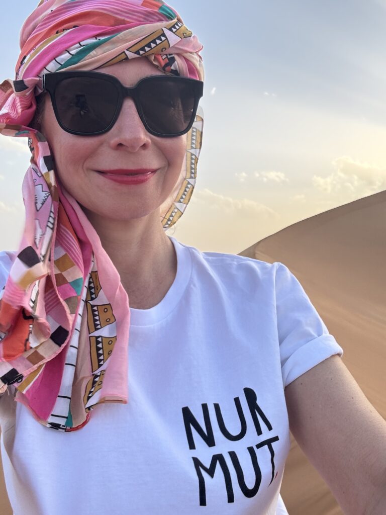 Jenny Heyer mit Sonnenbrille und gemustertem Kopftuch vor Sanddüne. Auf dem Shirt steht „NUR MUT“.