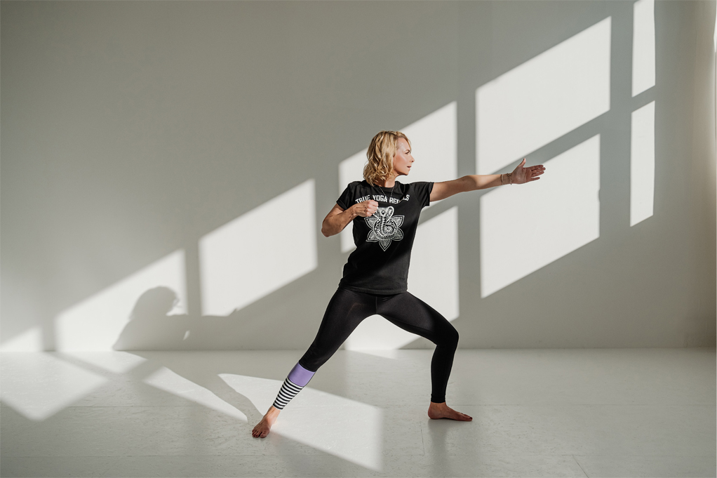 Jenny Heyer in schwarzer Kleidung in Yoga-Pose vor heller Wand mit rechteckigen Lichtflächen.